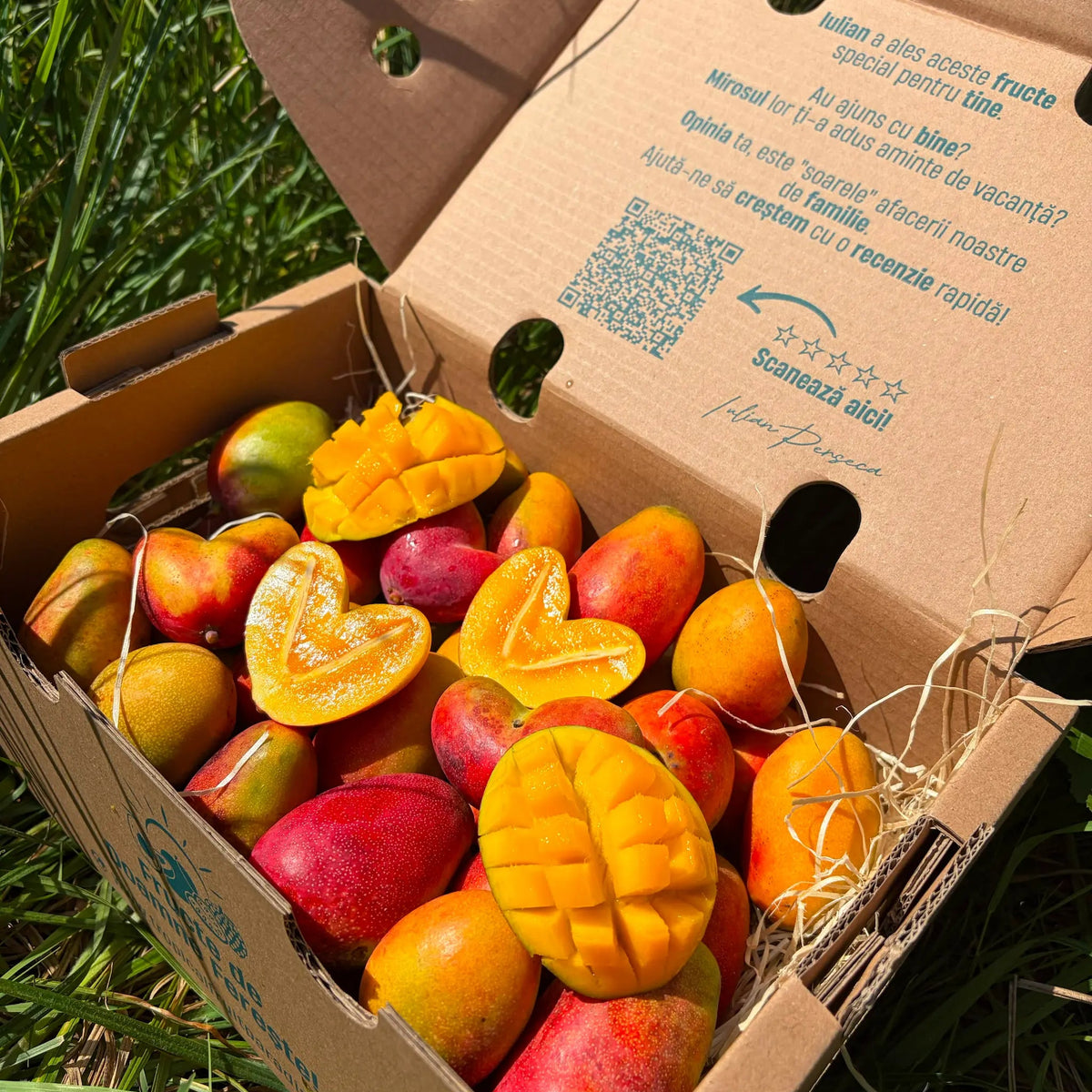Open cardboard delivery box filled with small red and orange Baby Irwin mangoes, featuring heart-shaped sliced halves and hedgehog cuts on a straw bed in green grass.