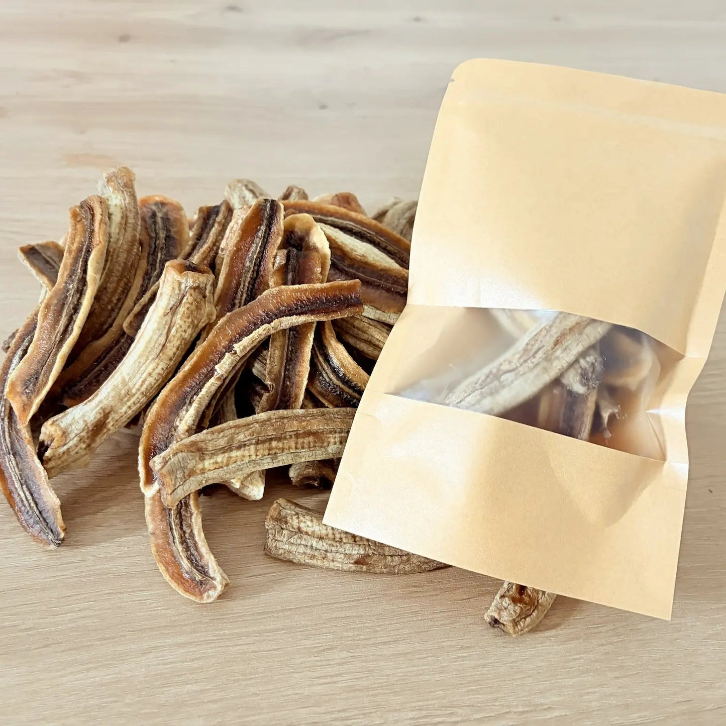 Natural dried banana strips with chewy texture, displayed next to an eco-friendly kraft paper pouch on a light wood background.