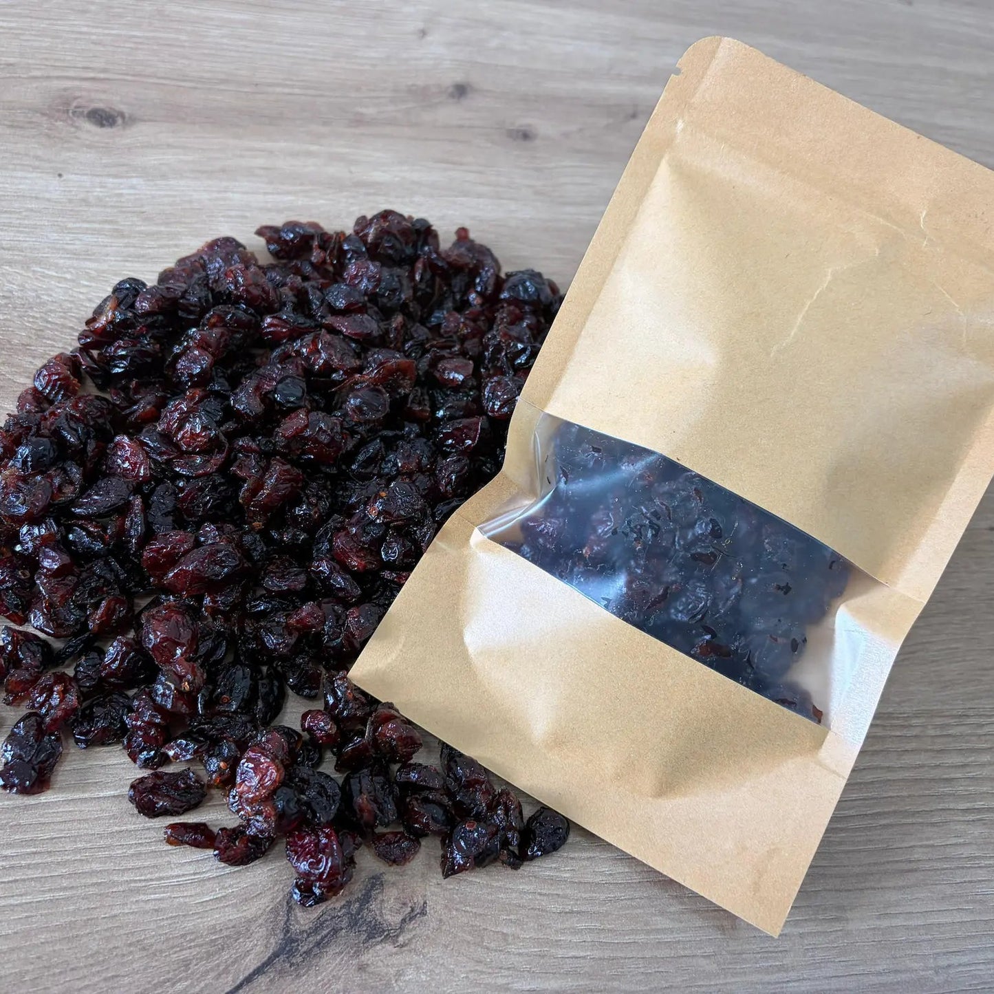 Natural dried cranberry with deep dark color and wrinkled texture, displayed next to an eco-friendly kraft paper pouch on a light wood background.