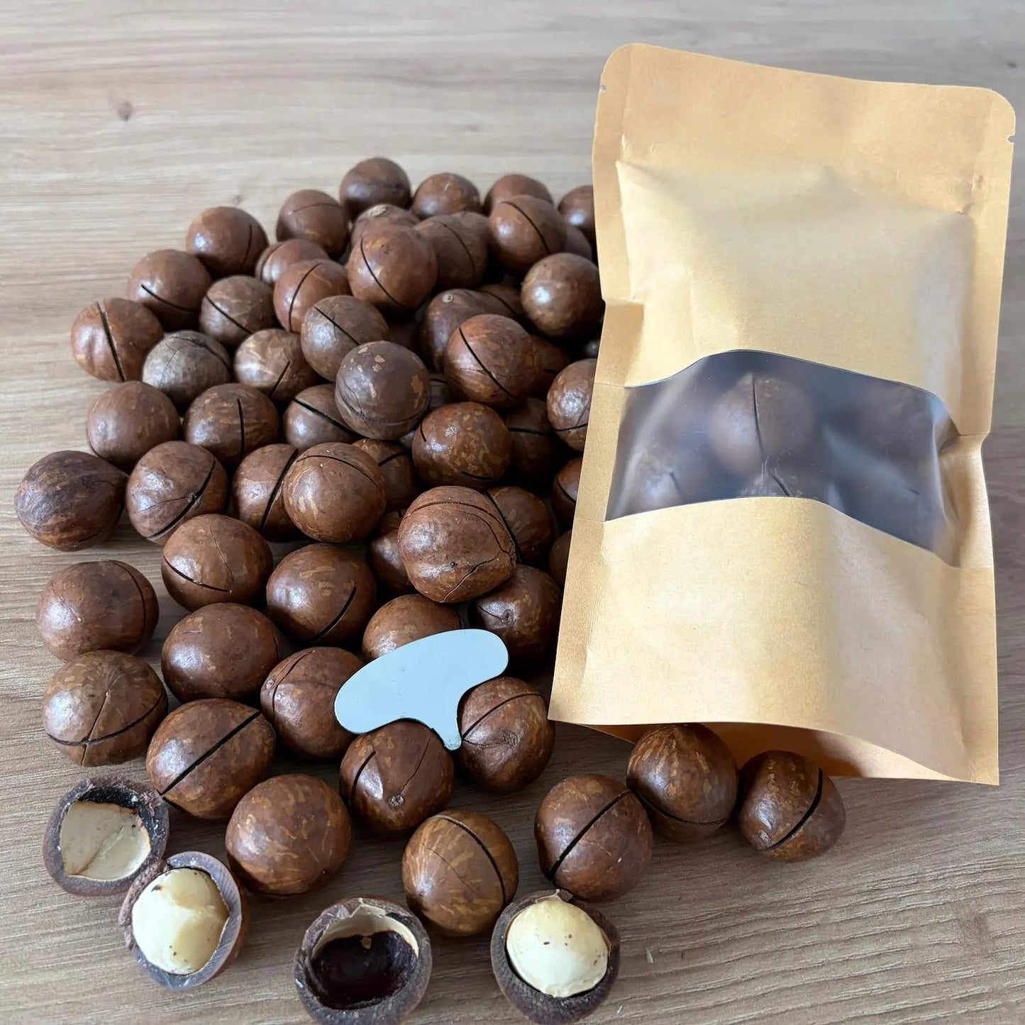 Premium whole macadamia nuts in shell with pre-cut slits, showing open creamy kernels and a metal key opener next to a kraft paper pouch on a wooden table.