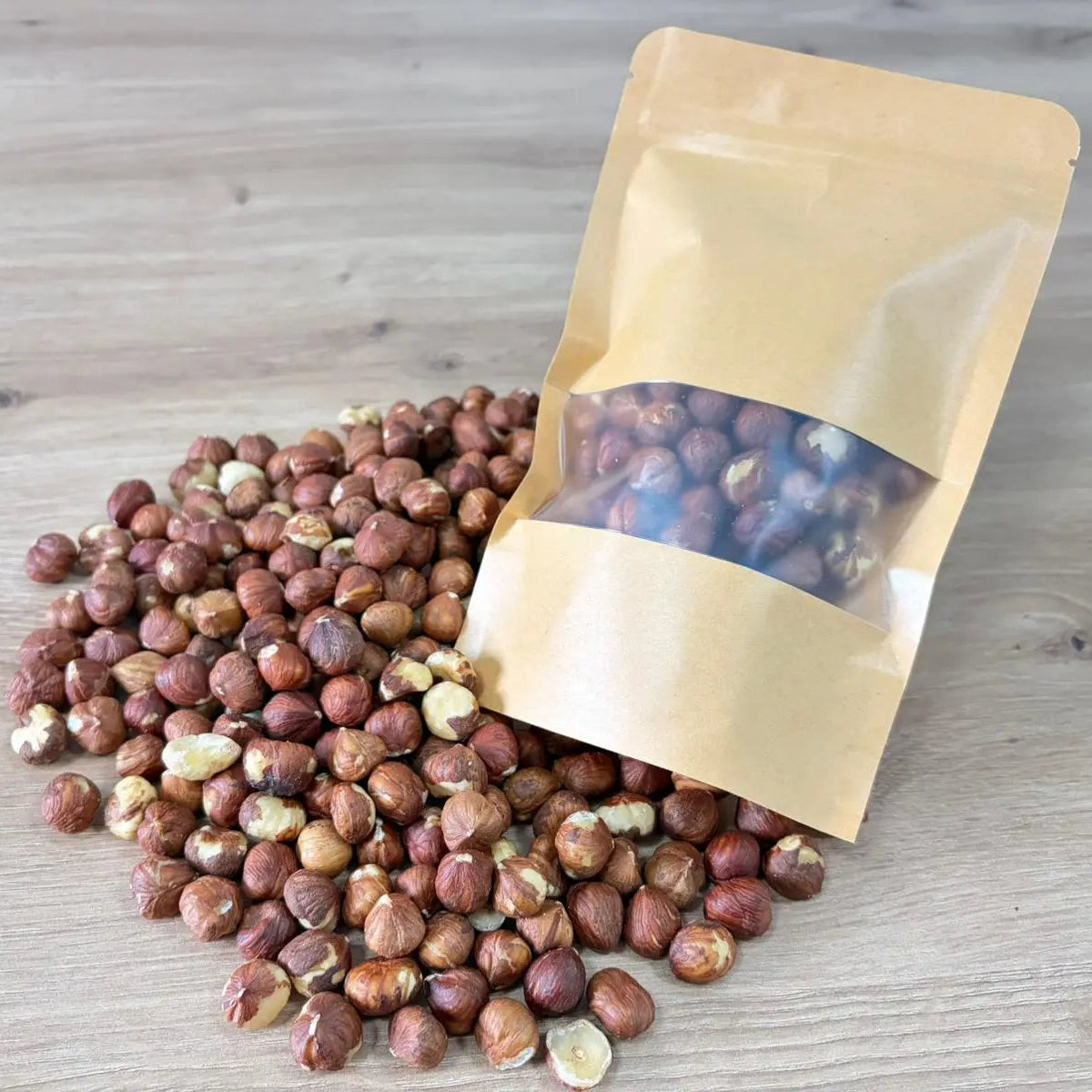 Raw hazelnut kernels with natural skin, displayed next to an eco-friendly kraft paper pouch on a light wooden surface.