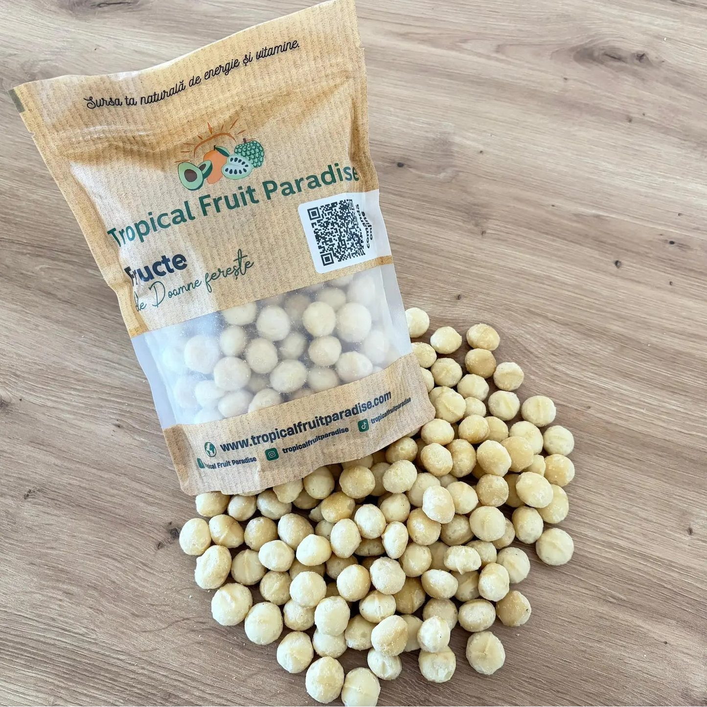 Raw shelled macadamia nut kernels spilled from a branded Tropical Fruit Paradise kraft pouch, displayed on a light wooden background.