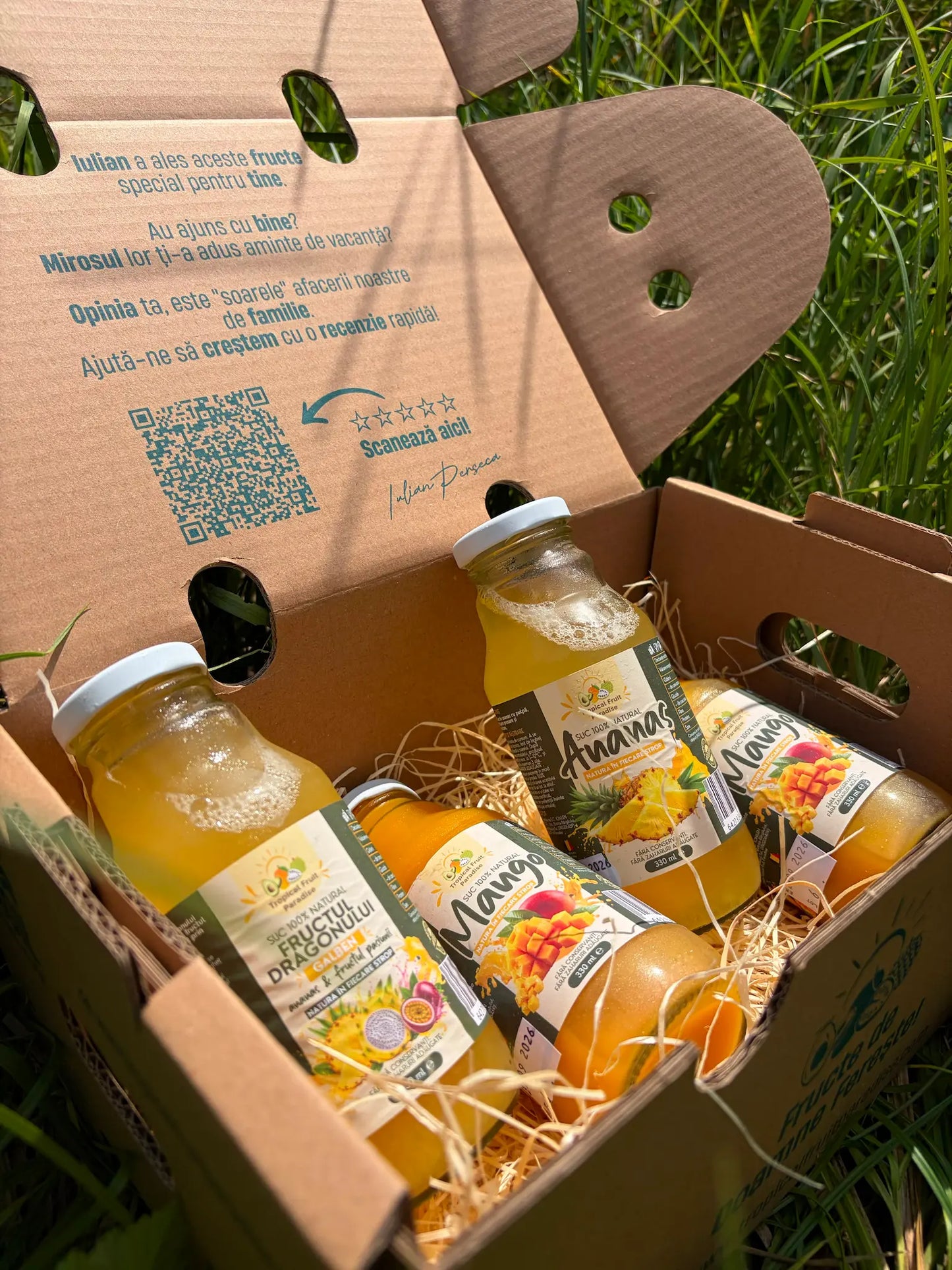 Four 330ml glass bottles of 100% natural tropical fruit juices (Mango, Pineapple, Yellow Dragon Fruit), resting on a straw bed inside an open cardboard box on green grass.