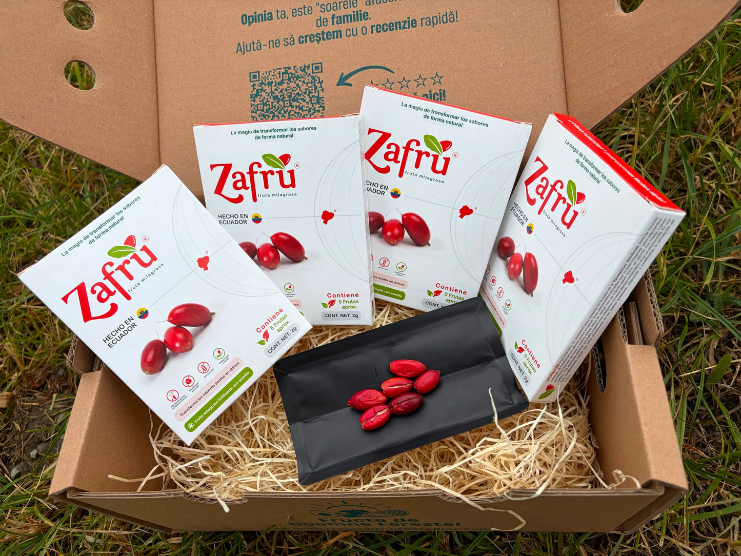 Zafru miracle berry (baya milagrosa) boxes with freeze-dried red berries displayed on an open black sachet, inside a cardboard delivery box on grass.
