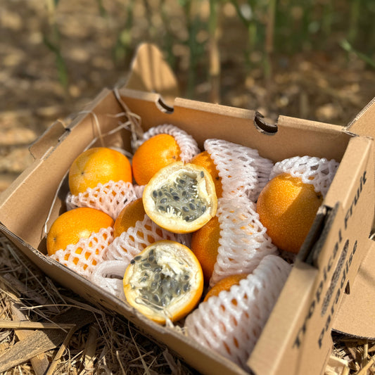 Box of granadilla with one cut open on a straw surface with plants in the background