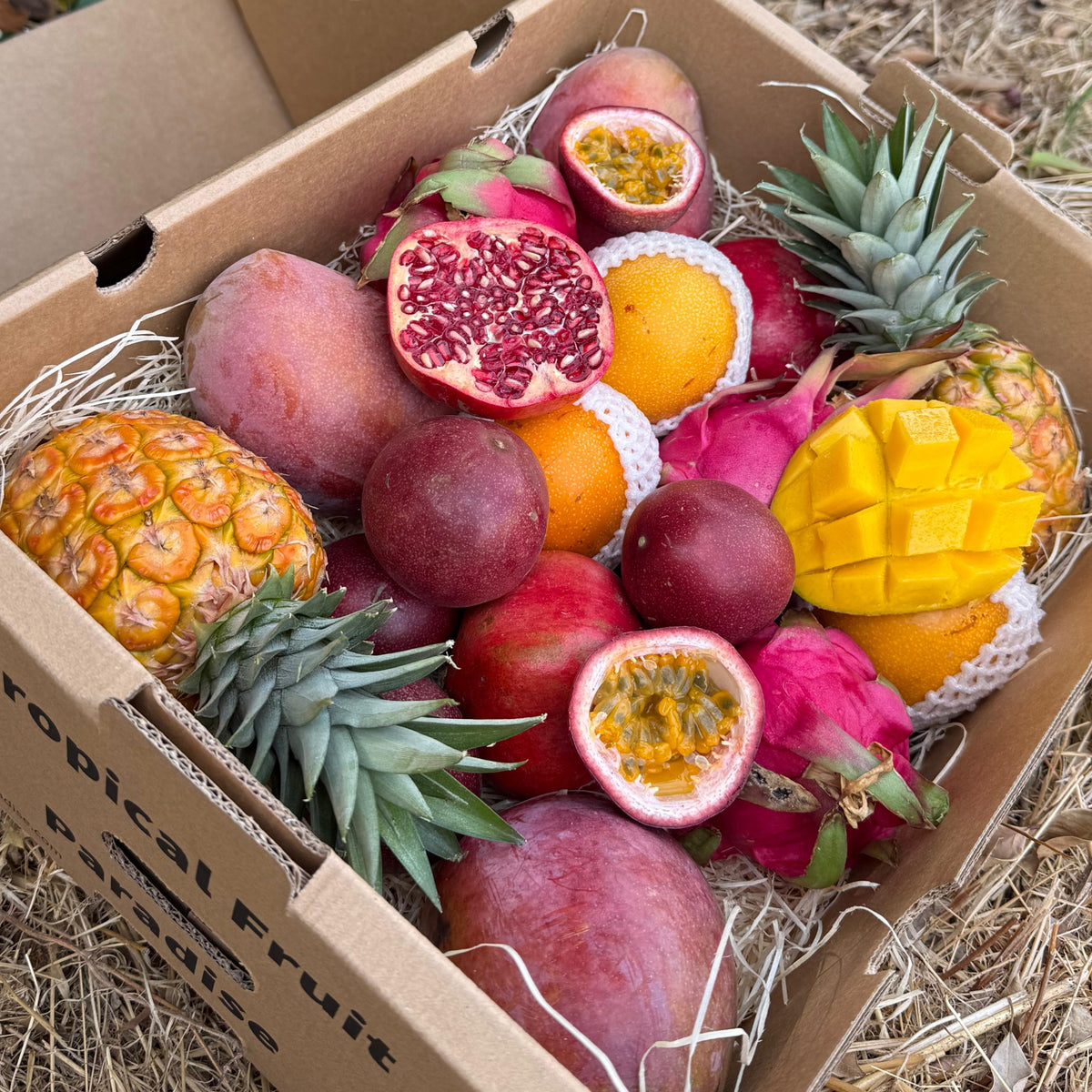 Box of assorted fruits including pineapples, mangoes, and pomegranates on a natural background.