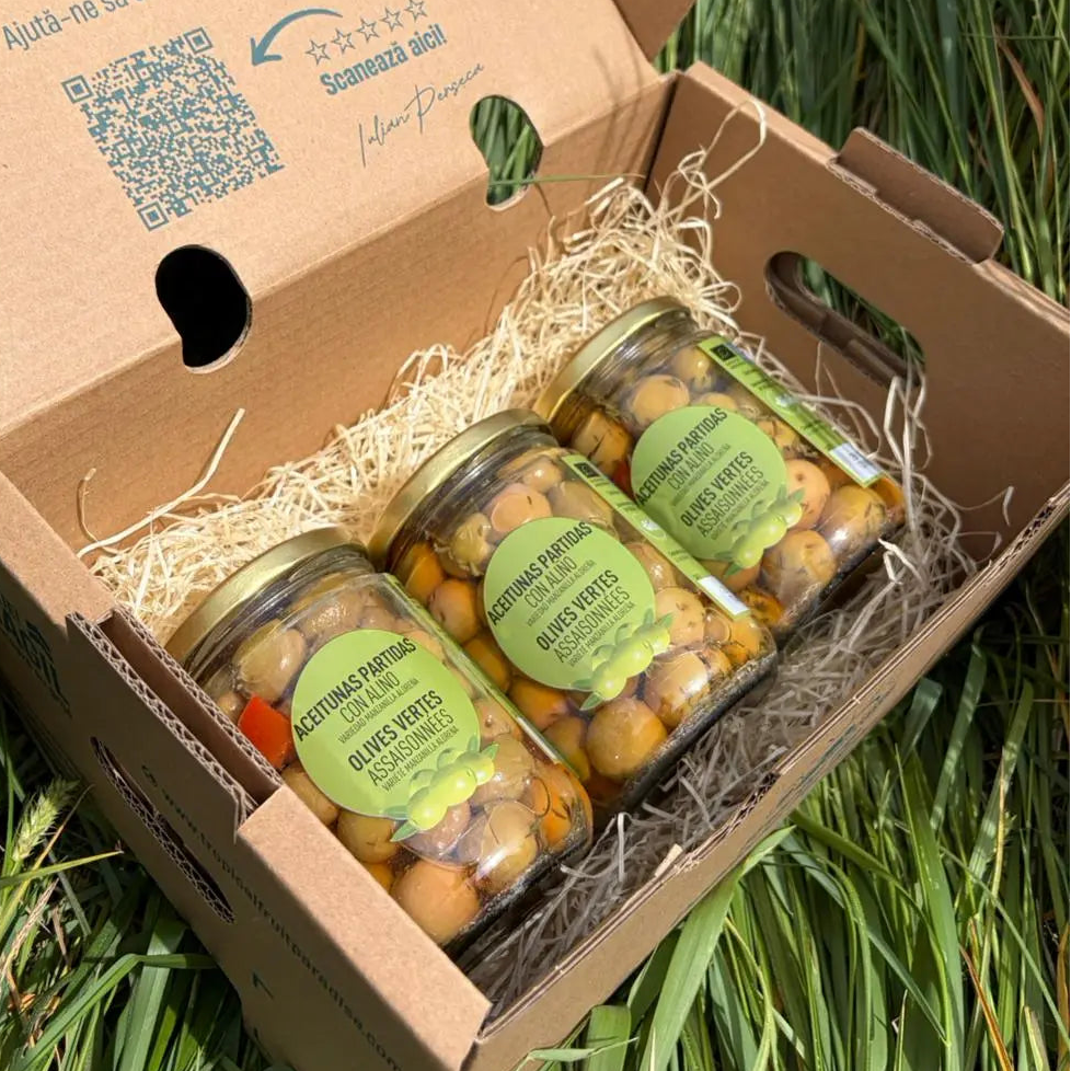 Three small 250g glass jars of seasoned cracked green olives (Aceitunas Partidas con Aliño), neatly arranged on straw inside an open cardboard box on green grass.