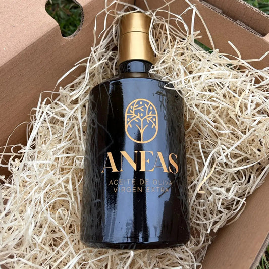 Premium black bottle of ANEAS extra virgin olive oil with gold lettering, nestled in protective straw inside a cardboard delivery box on green grass.