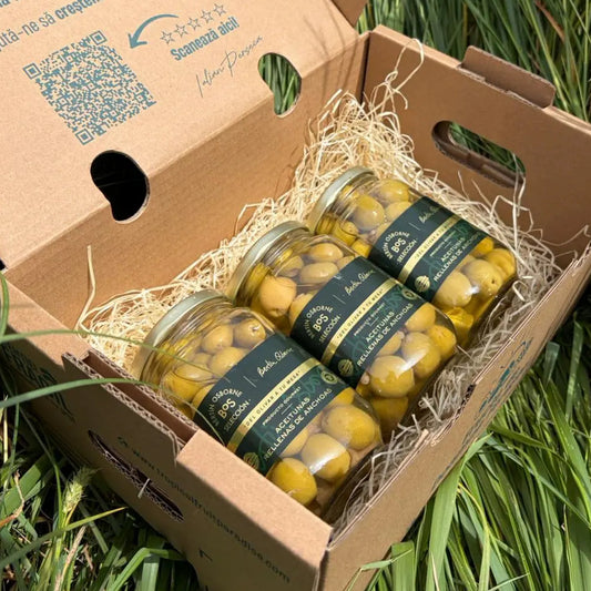 Three 180g glass jars of premium green Manzanilla olives stuffed with anchovy paste from the BOS Selección range, resting on a straw bed inside an open cardboard box on grass.