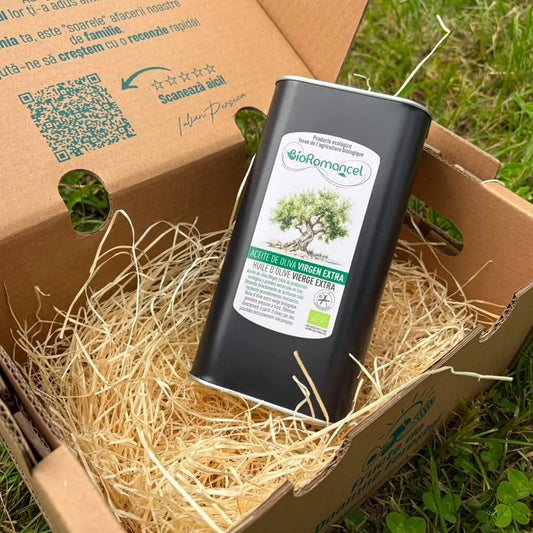 Black 1-liter metal tin of BioRomancel organic extra virgin olive oil, resting on a bed of straw inside an open cardboard delivery box on green grass.