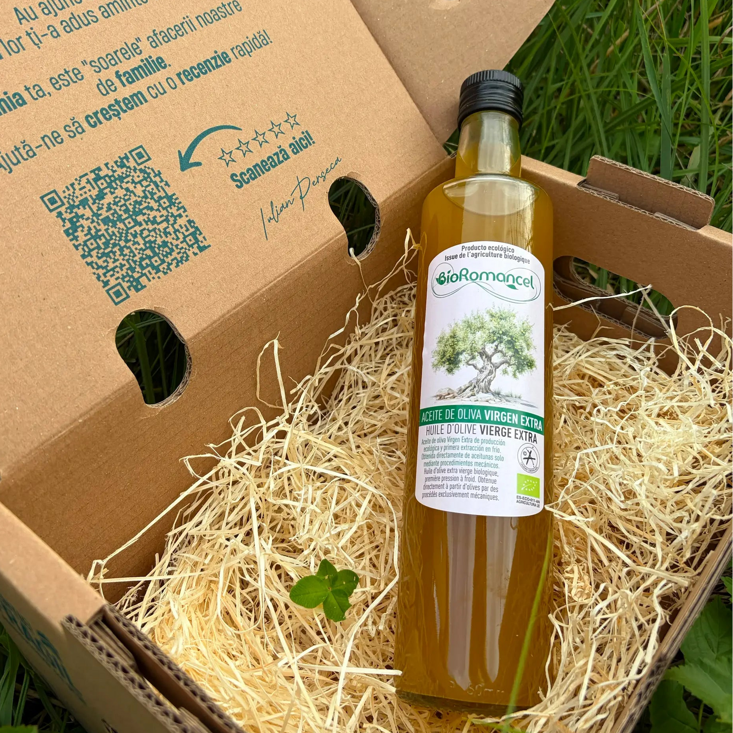 A clear 500ml glass bottle of BioRomancel organic extra virgin olive oil, showing its golden color, resting on a bed of straw inside a delivery box with a four-leaf clover, on grass.