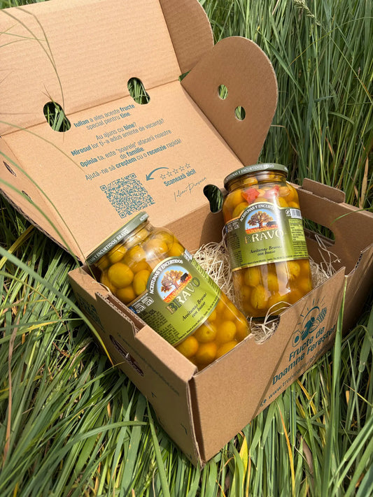 Three 525g glass jars of spicy Bravo Aceitunas Bravas olives with pits, neatly arranged on a straw bed inside an open cardboard delivery box on green grass.