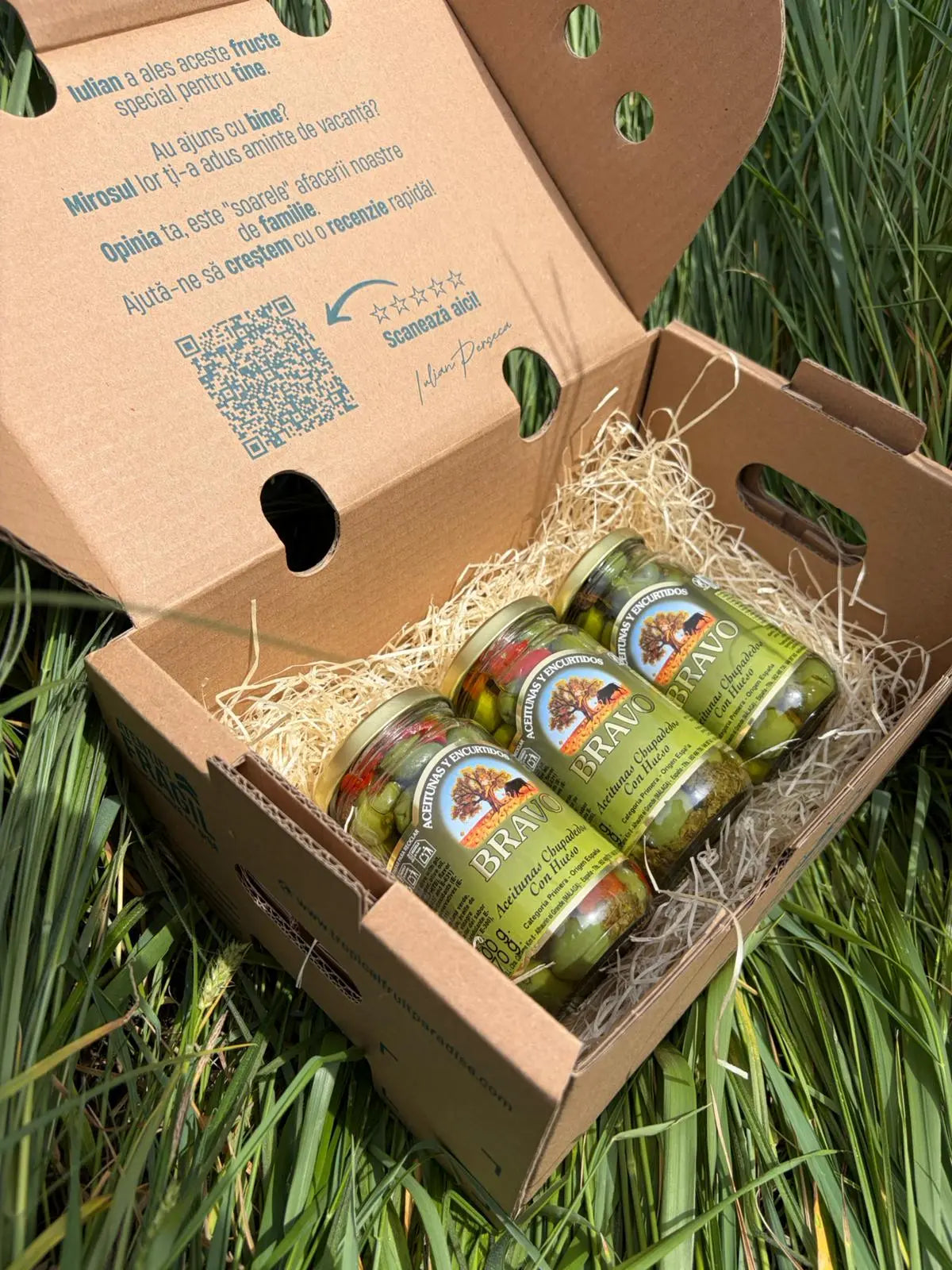 Three small 170g glass jars of green Bravo Aceitunas Chupapedos olives with pits, arranged on a straw bed inside an open cardboard box on green grass.