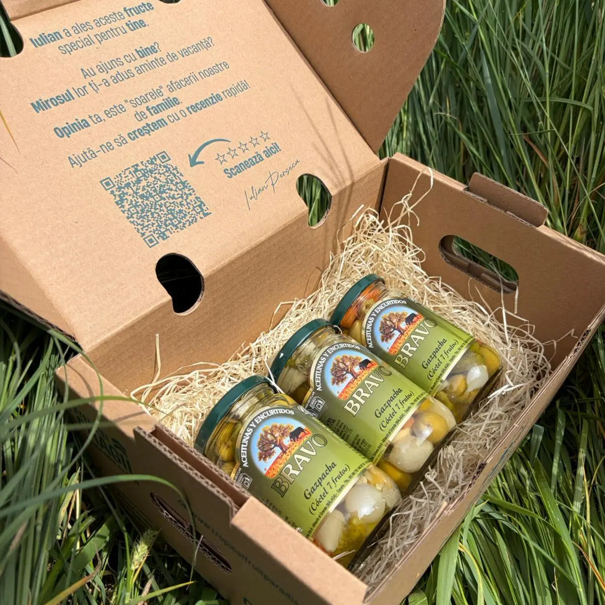 Three small 170g glass jars of Bravo Gazpacha mixed olives and pickled vegetables (Cóctel 7 frutos), arranged on a straw bed inside a cardboard box on green grass.