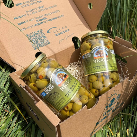 Two large 500g glass jars of giant green Gordal olives stuffed with red pepper paste (Aceitunas Gordal rellenas de pimiento) by Bravo, resting on straw inside a cardboard delivery box on green grass.