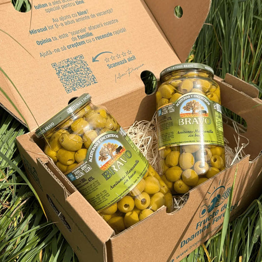 Two large 475g glass jars of pitted green Manzanilla olives by Bravo, highlighting the empty centers of the olives, resting on straw inside a cardboard box on grass.