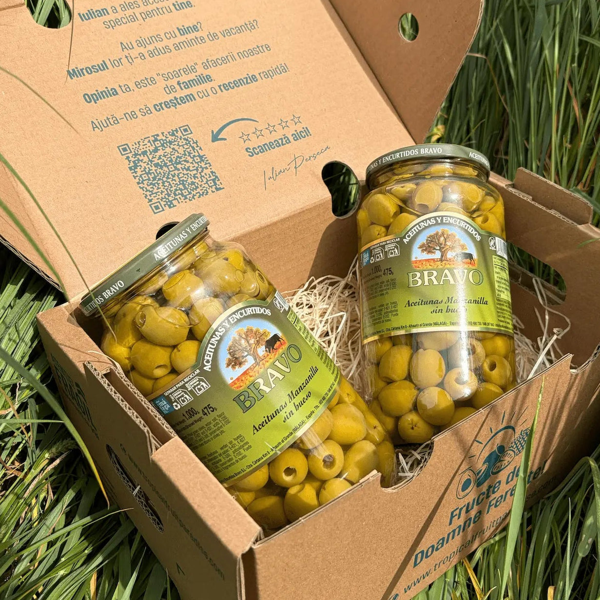 Two large 475g glass jars of pitted green Manzanilla olives by Bravo, highlighting the empty centers of the olives, resting on straw inside a cardboard box on grass.