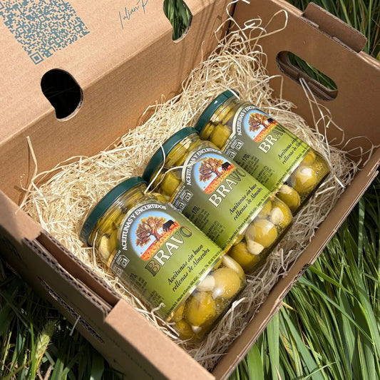 Three small 170g glass jars of green Bravo olives stuffed with crunchy almonds (Aceitunas rellenas de almendra), arranged on straw inside a cardboard delivery box on green grass.