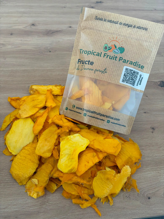 Large golden-orange dehydrated mango slices spilling out of a branded Tropical Fruit Paradise kraft pouch onto a wooden table.