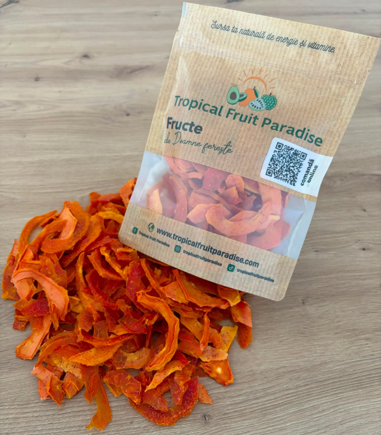 Vibrant reddish-orange dehydrated papaya strips spilling out of a branded Tropical Fruit Paradise kraft pouch onto a wooden table.