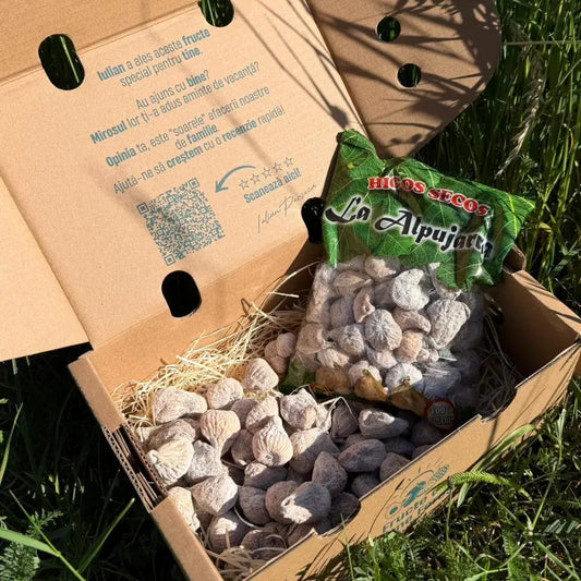 Natural dried figs coated in flour, displayed loose and in their original Higos Secos La Alpujarra bag, resting on straw inside a delivery box on green grass.