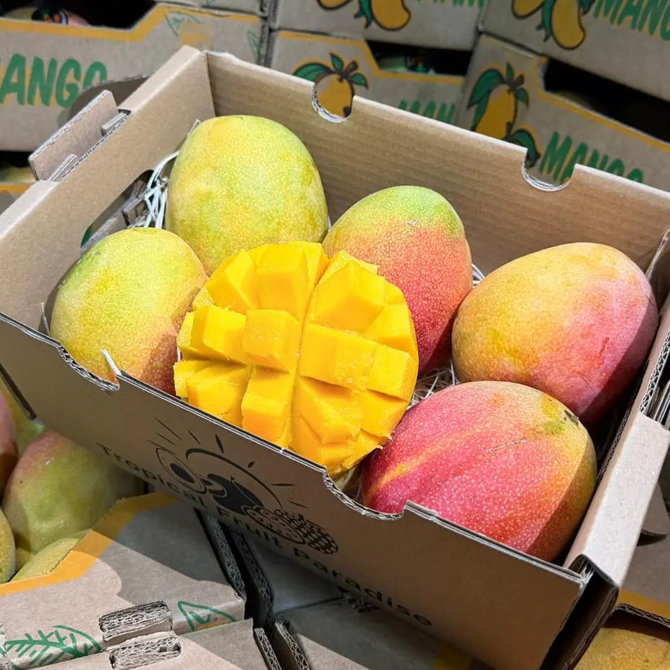 Fresh Haden mangoes in a delivery box, with one fruit sliced in a hedgehog pattern to show the juicy yellow flesh, against a background of stacked mango crates.