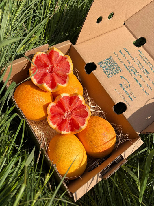 Premium red grapefruit, whole and decoratively star-cut to show the juicy ruby red flesh, resting on straw inside an open delivery box on green grass.