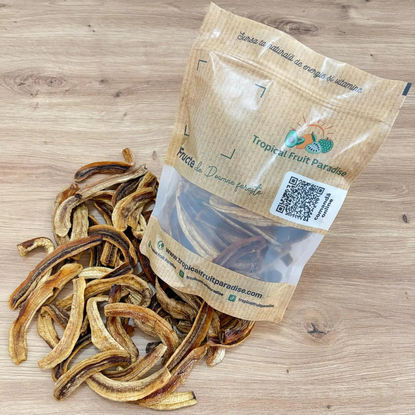Natural dried banana strips with caramelized golden tones, spilled from a 500g branded Tropical Fruit Paradise kraft pouch, displayed on a light wooden table.
