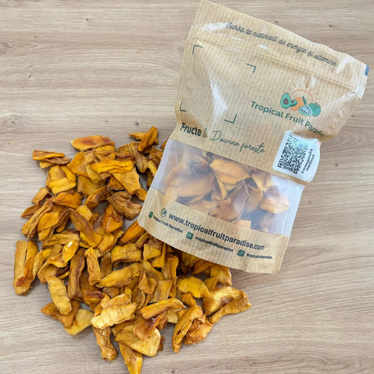 Natural dried mango slices with a vibrant golden-yellow color, spilled from a 500g branded Tropical Fruit Paradise kraft pouch, displayed on a light wooden table.