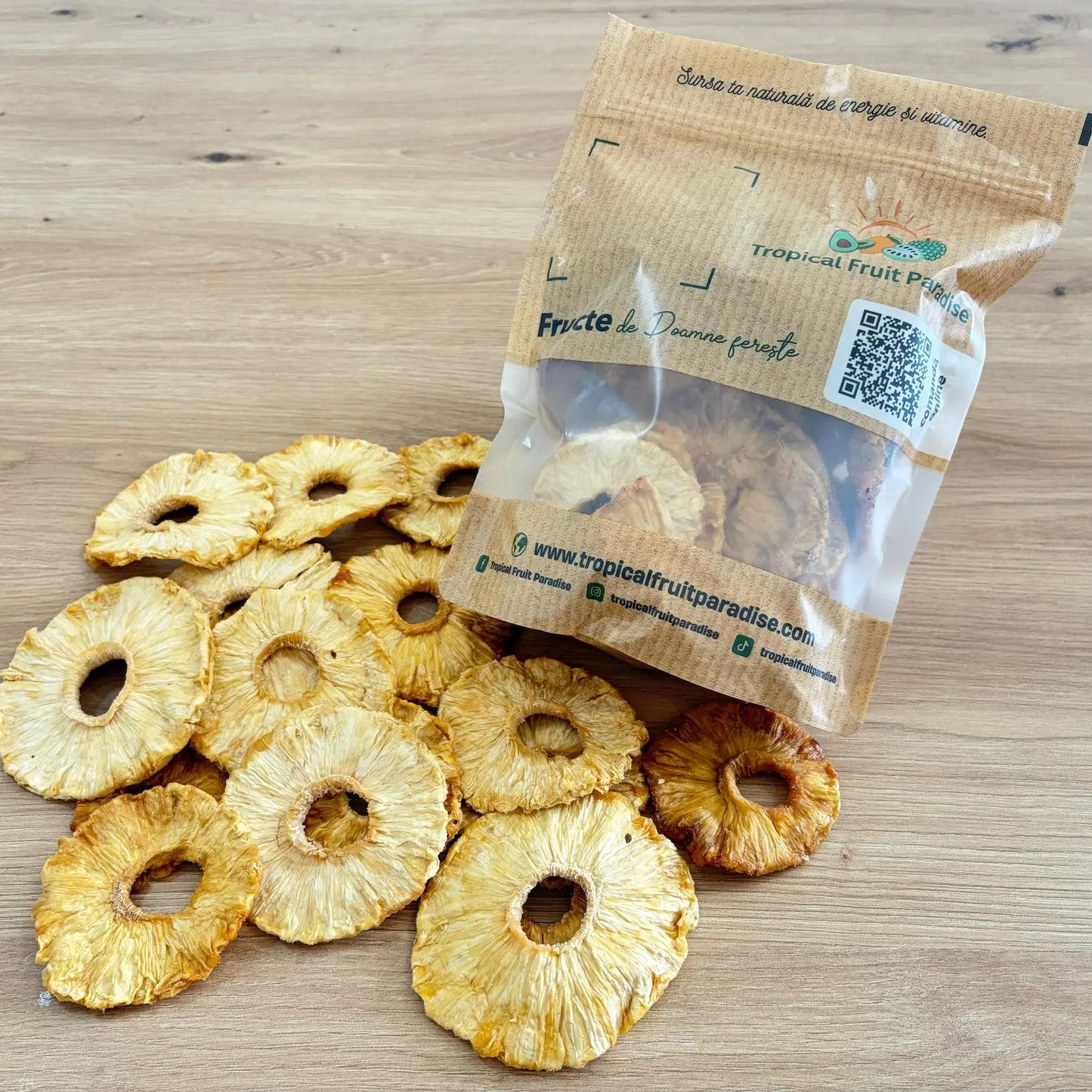 Natural dried pineapple rings with a golden yellow color, spilled from a 500g branded Tropical Fruit Paradise kraft pouch, displayed on a light wooden table.