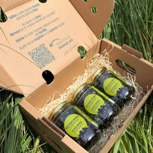 Three small 250g glass jars of organic dried black olives (Aceitunas Negras Deshidratadas), arranged on straw inside a cardboard delivery box on green grass.