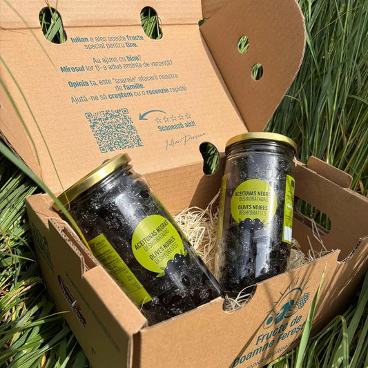 Two large 500g glass jars of organic dried black olives, displaying the eco certification on the label, resting on a bed of straw inside a delivery box on grass.