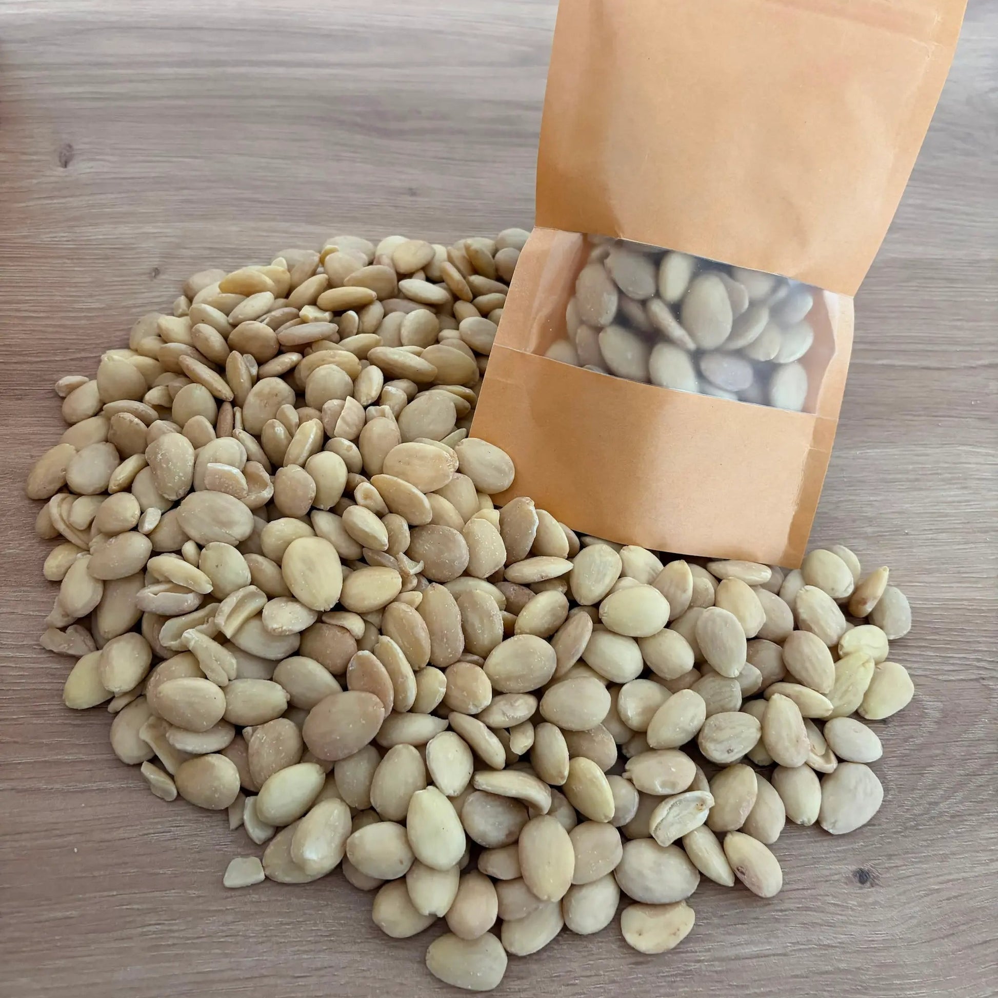 Premium peeled Belona almonds with a round, flat shape and light cream color, spilling out from a small kraft pouch onto a wooden table.