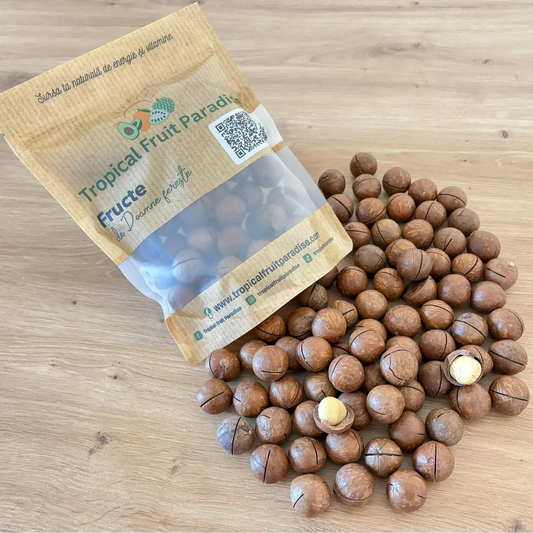 Premium whole macadamia nuts in shell with pre-cut slits, displaying open creamy white kernels, spilled from a 500g branded Tropical Fruit Paradise kraft pouch on a light wooden table.