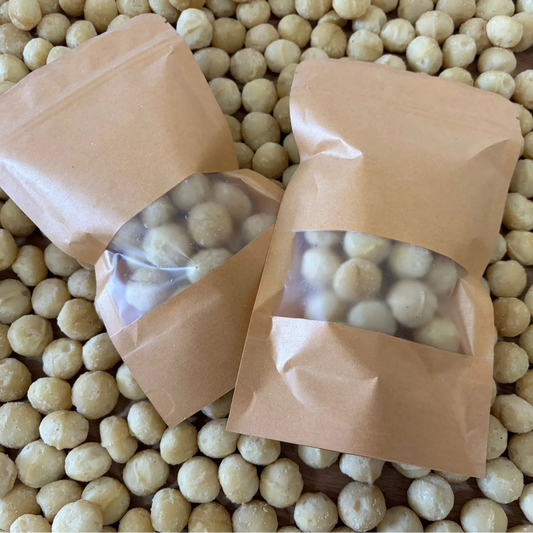 Raw crunchy macadamia nuts in eco-friendly kraft paper pouches, displayed on a background of premium macadamia nut kernels.