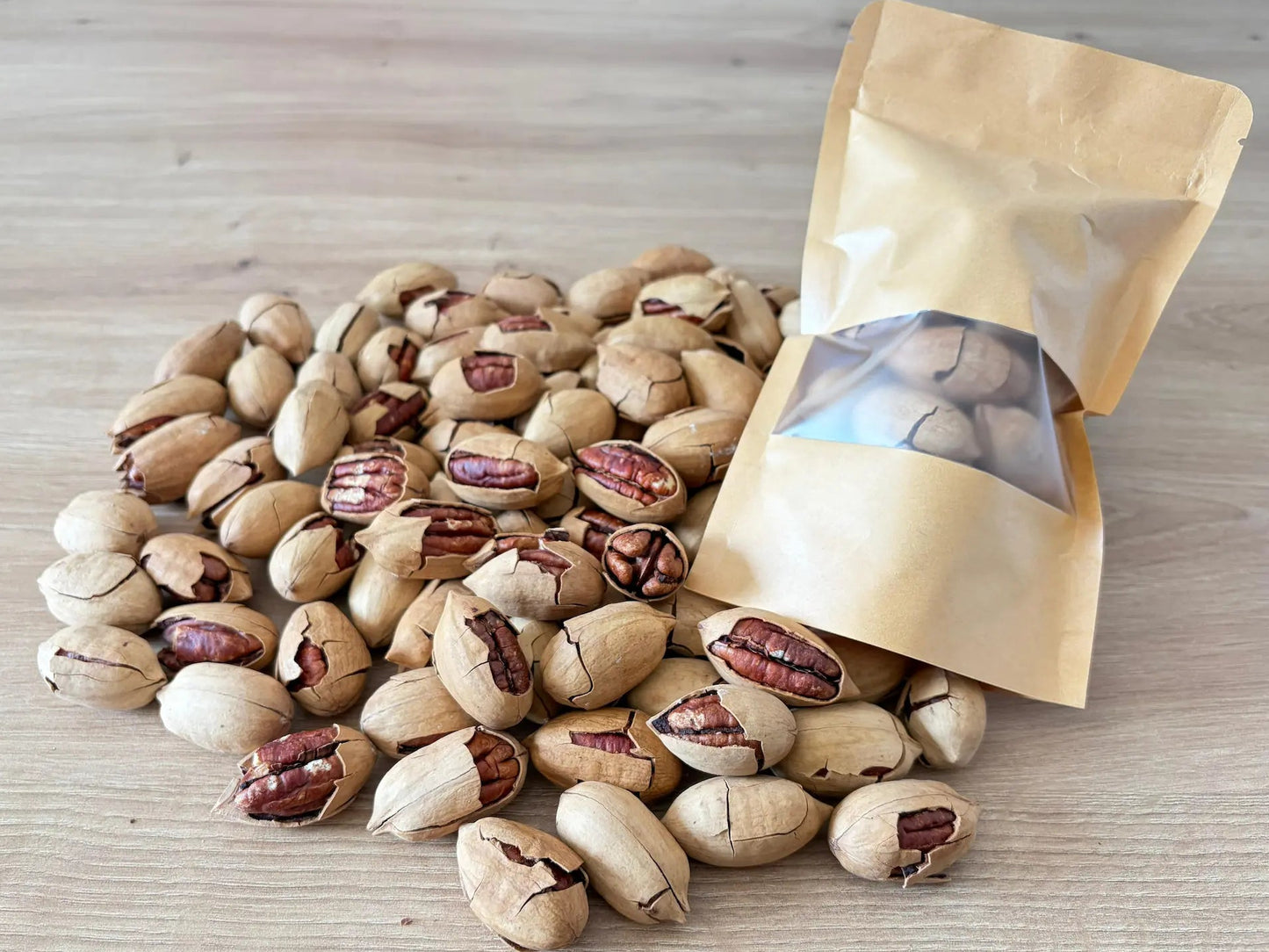 Raw pecan nuts in shell with pre-cracked hulls showing the wrinkled brown kernels, displayed next to a kraft paper pouch on a light wooden surface.