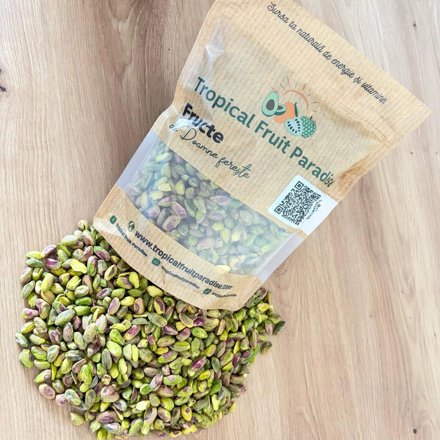 Raw shelled pistachio kernels with vibrant green and purple tones, spilled from a 500g branded Tropical Fruit Paradise kraft pouch, displayed on a light wooden table.