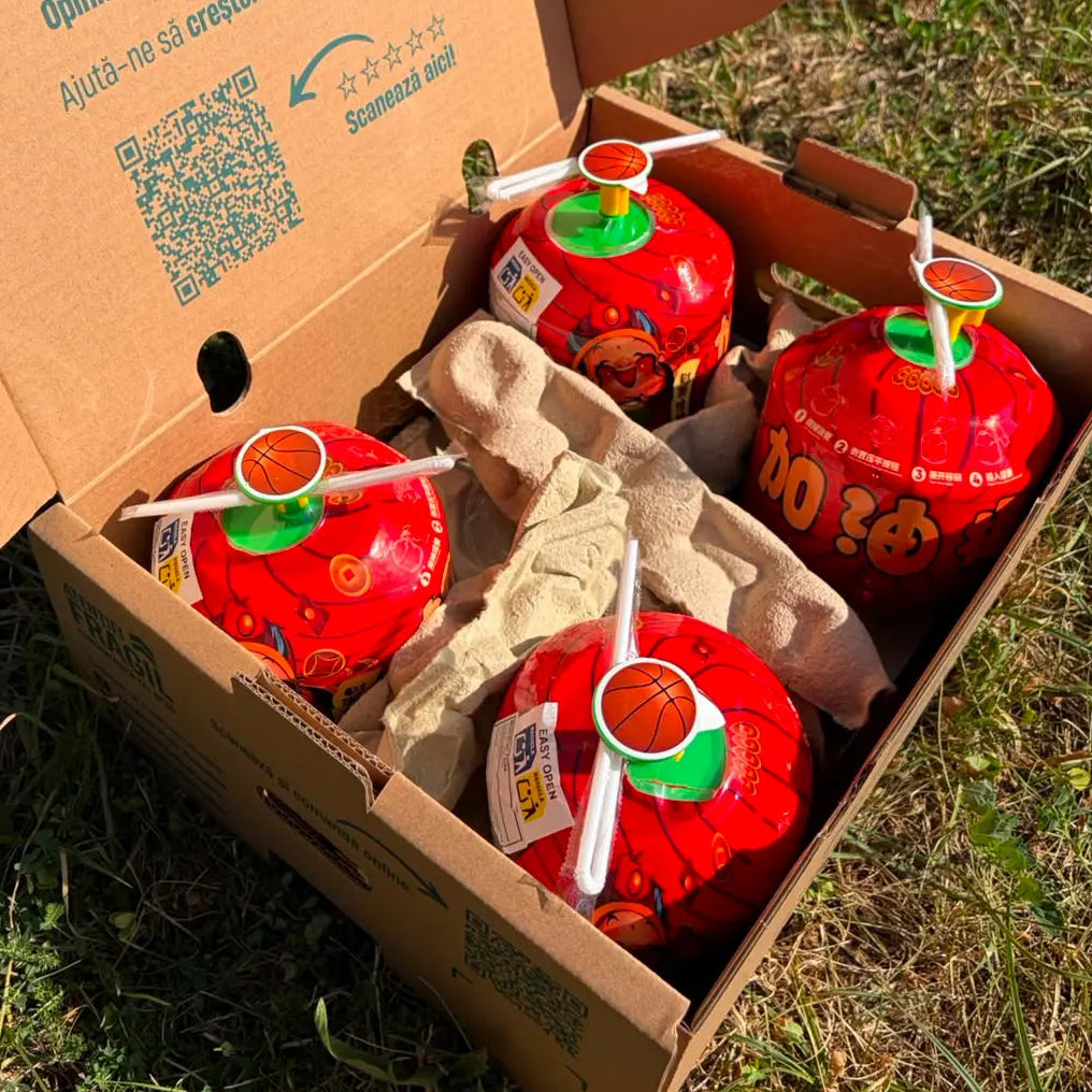 Open delivery box containing four ready-to-drink red coconuts with easy-open tabs and straws, resting on a protective cardboard insert on green grass.