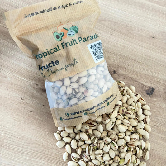 Roasted in-shell pistachios with partially opened naturally cream hulls showing the green-golden kernels, spilled from a branded Tropical Fruit Paradise kraft pouch on a light wooden table.