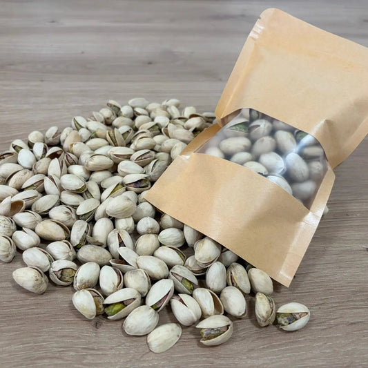 Roasted and salted pistachios in shell, displayed next to an eco-friendly kraft paper pouch on a light wood background.