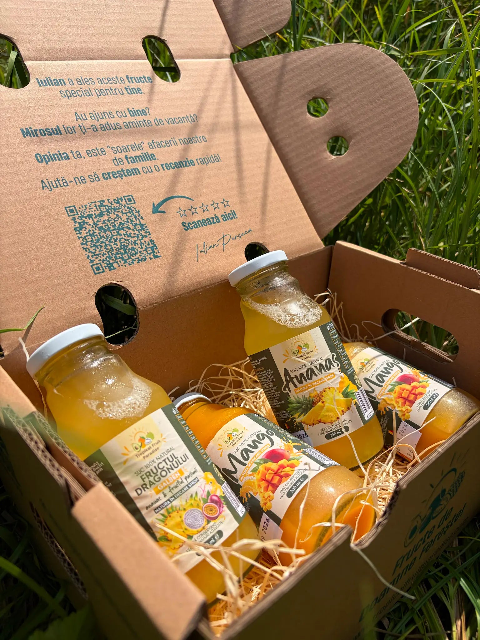 Four 330ml glass bottles of 100% natural tropical fruit juices (Mango, Pineapple, Yellow Dragon Fruit), resting on a straw bed inside an open cardboard box on green grass.