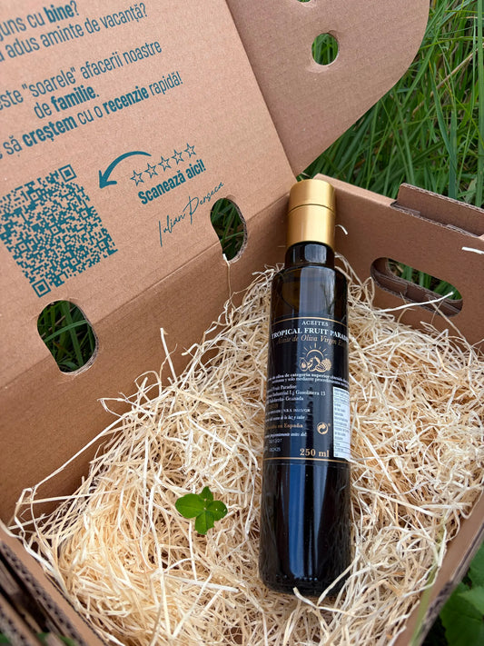 An elegant 250ml glass bottle of Tropical Fruit Paradise extra virgin olive oil with a gold cap, resting on straw inside a cardboard box with a lucky four-leaf clover, on green grass.