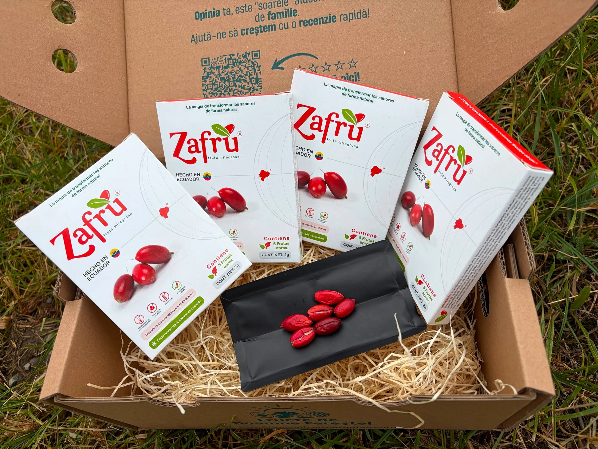 Zafru miracle berry (baya milagrosa) boxes with freeze-dried red berries displayed on an open black sachet, inside a cardboard delivery box on grass.